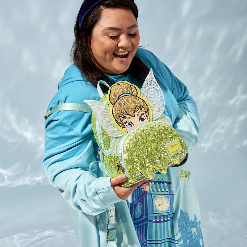 Woman wearing a blue Peter Pan Loungefly hoodie and holding the green and gold Loungefly Peter Pan Tinker Bell Exclusive Sequin Cosplay Mini Backpack against a shimmery sky background